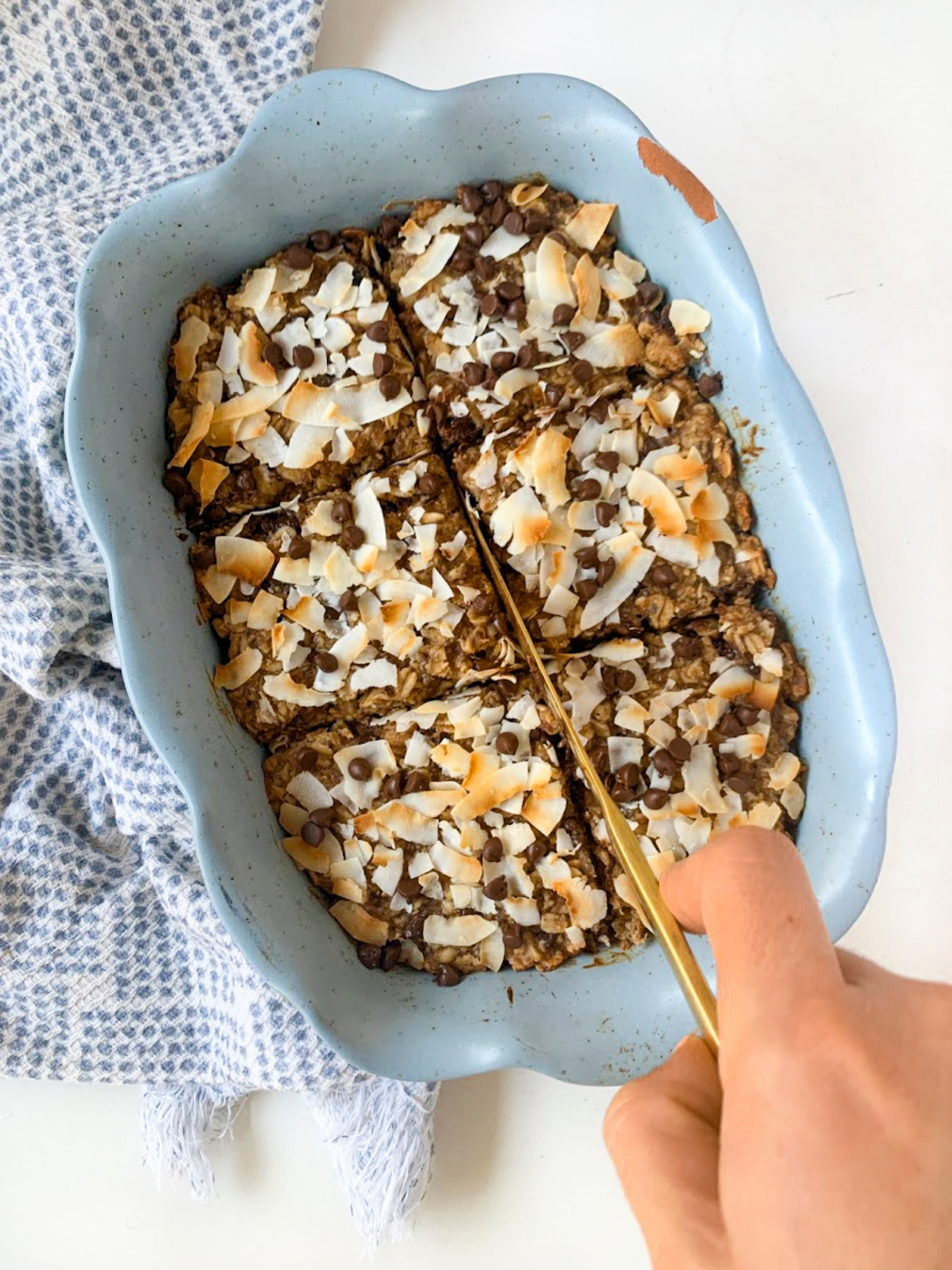 cutting into baked oatmeal topped with chocolate chips and coconut flakes. Sitting on white table with blue kitchen towel